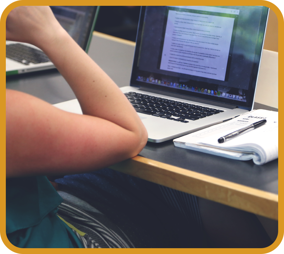 Someone seated by a desk with a laptop, notebook and pen